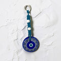 Mati Made Azure Disc wall charm featuring a blue acrylic evil eye pendant with braided cord and white accent beads.