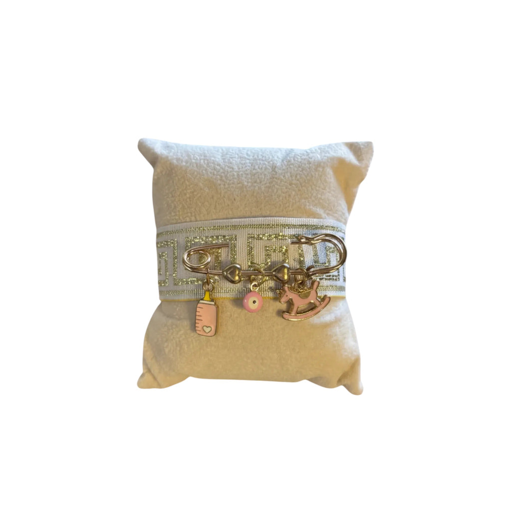 Mati Made baby keepsake pin with gold ribbon, evil eye, baby bottle, and rocking horse charms on a display pillow.
