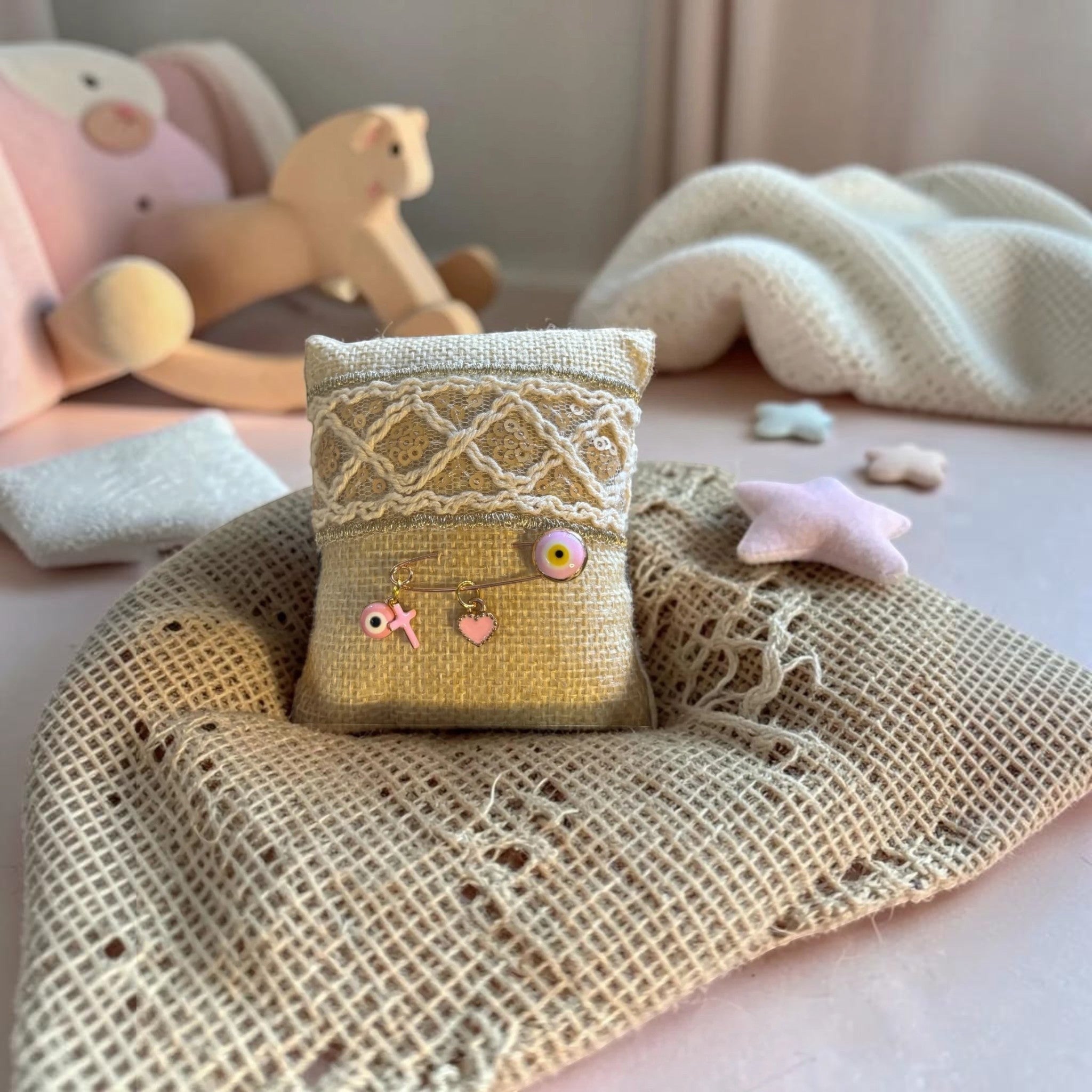 Mati Made gold-tone baby keepsake pin with pink evil eye, cross, and heart charms on a decorative textured cushion.