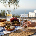 Mati Made clear plastic can-style tumbler with blue eye pattern, lid, and straw on a Mediterranean picnic table.