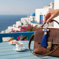 Mati Made beige faux-leather cross keychain with evil eye charm and blue silk tassel attached to a brown leather bag.