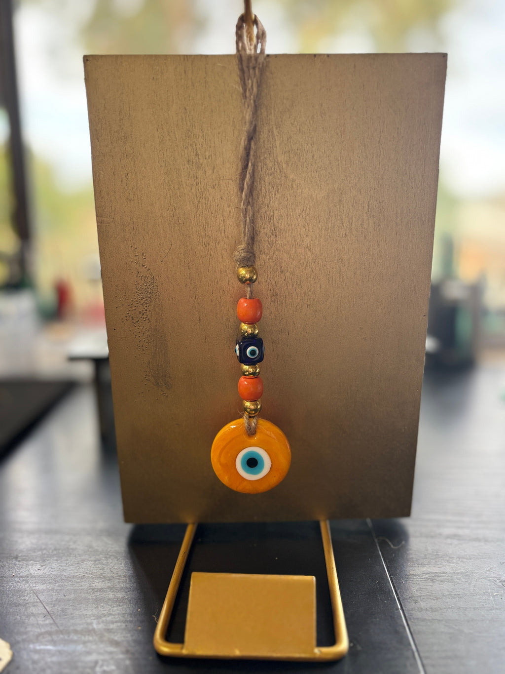 Mati Made orange and blue evil eye glass charm hanging on a jute string with decorative beads on a gold stand