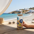 Mati Made glass tumbler with evil eye motif, bamboo lid, and glass straw, held on a beachside table with iced coffee.