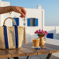 Mati Made evil eye keychain with a neutral faux leather tassel and silver hardware hanging over a straw tote bag