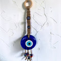 Mati Made handcrafted wall hanging with a blue evil eye, jute macrame cord, and light wooden ring on a white wall.