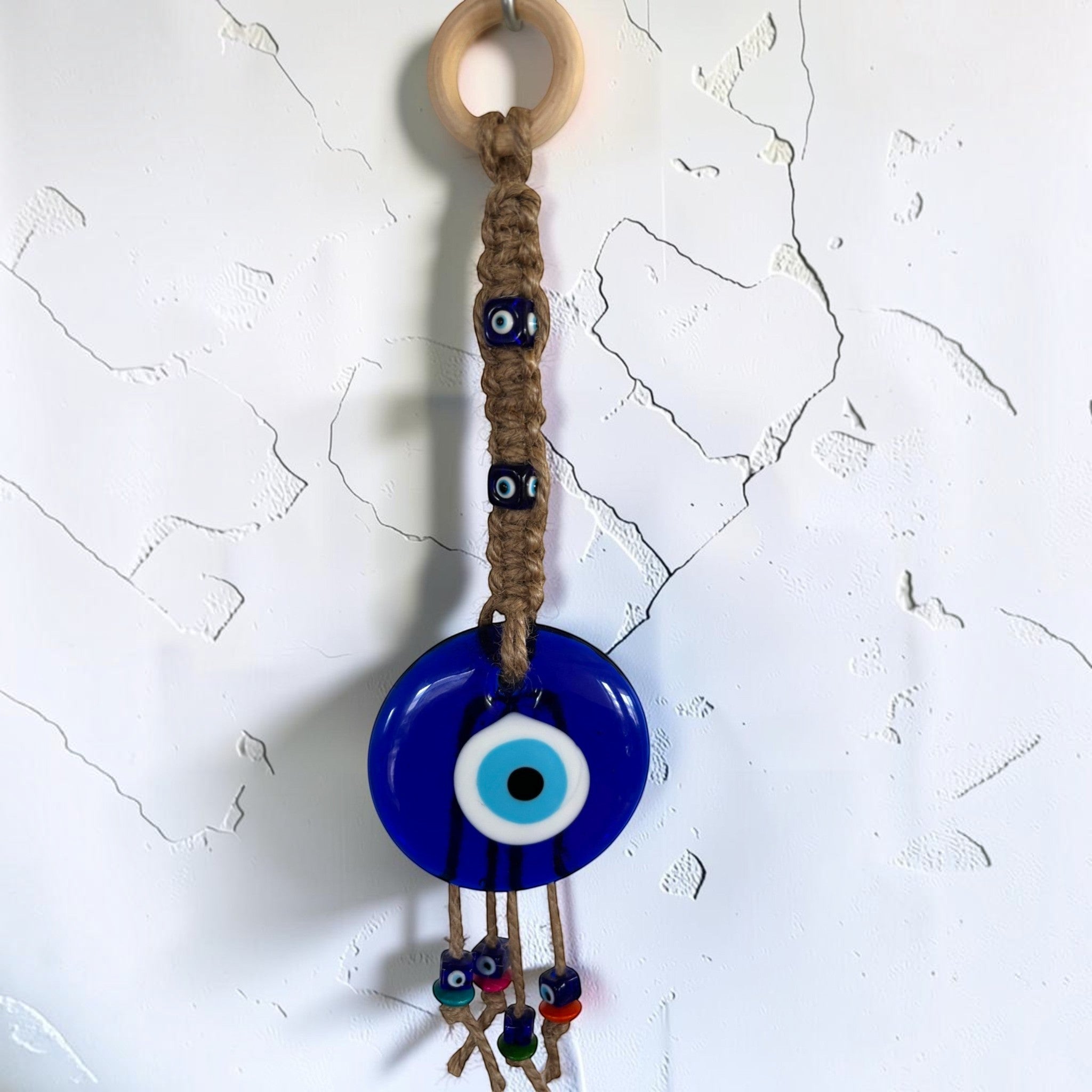 Mati Made handcrafted wall hanging with a blue evil eye, jute macrame cord, and light wooden ring on a white wall.
