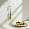 Mati Made evil eye wall hanging ornament with yellow and blue beads on a jute cord, displayed on a white textured wall.