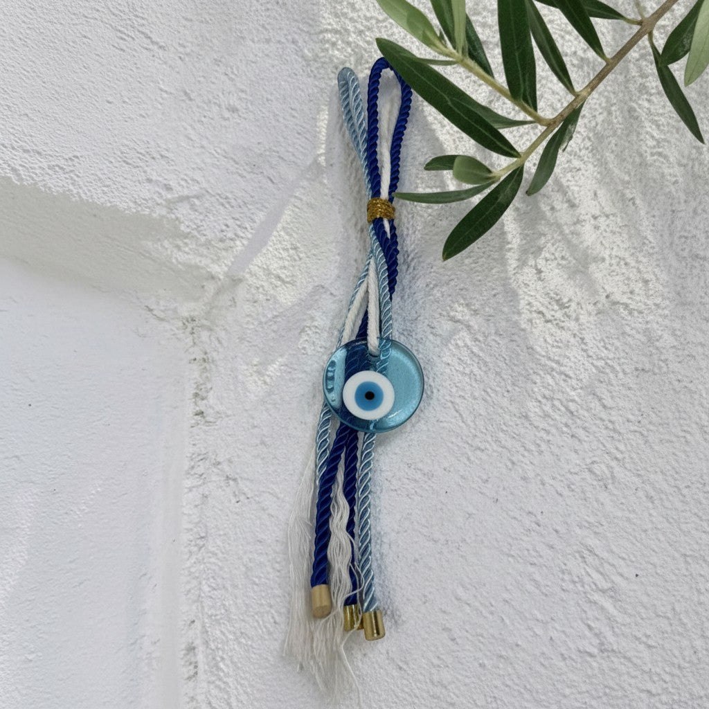 Mati Made light blue glass evil eye wall charm with twisted blue and white cords and gold accents on white wall