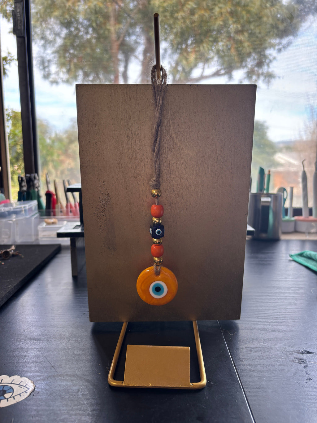 Mati Made orange evil eye glass charm with beads on a jute string hanging from a gold display stand