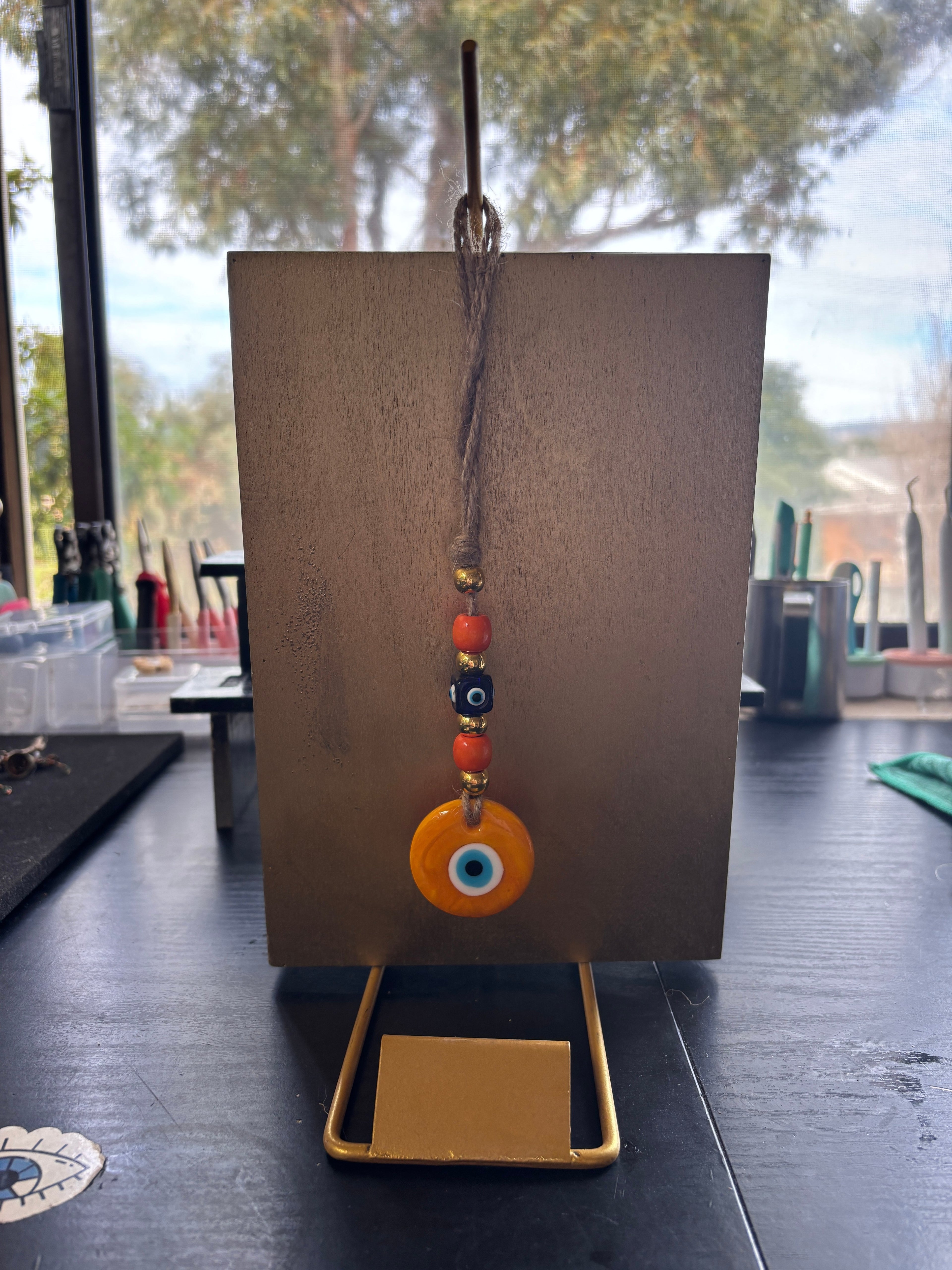 Mati Made orange evil eye glass charm with beads on a jute string hanging from a gold display stand
