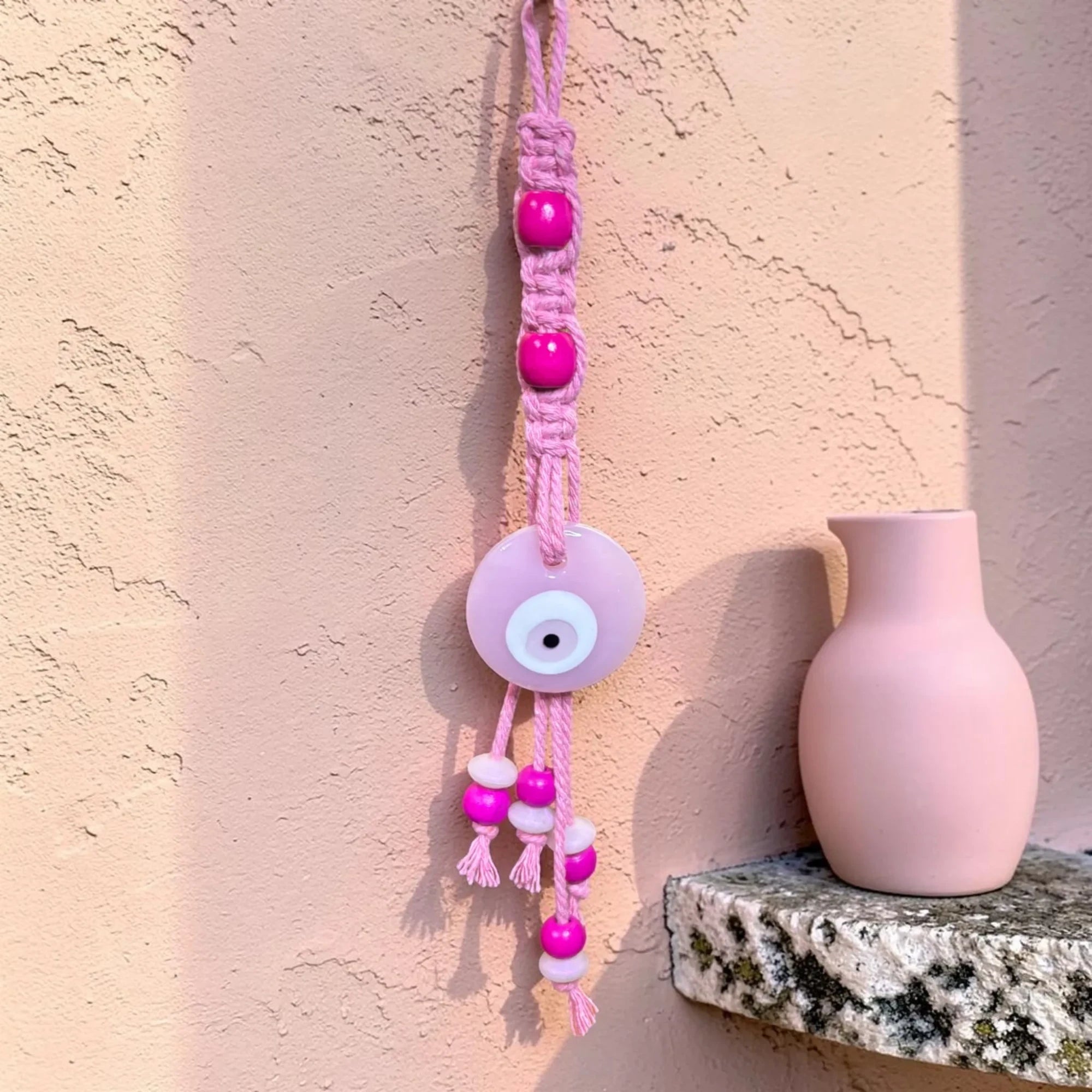 Mati Made pink macrame evil eye wall hanging with fuchsia wooden beads and a central glass charm on a textured wall.
