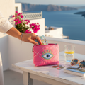 Mati Made hot pink quilted pouch with an evil-eye motif and sequin details, held on a white table overlooking the sea.
