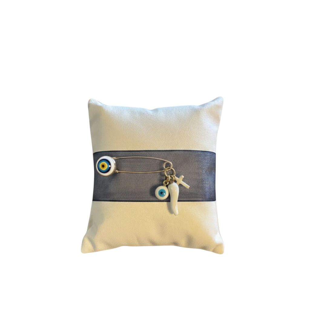 Mati Made protective baby nursery pin with blue evil eye, cross, and horn charms on a cream velvet cushion