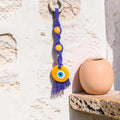 Mati Made handcrafted purple macrame evil eye wall hanging with yellow wooden beads and a glass charm on a stone wall.