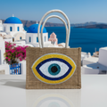 Mati Made natural jute tote bag featuring a hand-embellished sequin and bead evil eye design against a Greek backdrop.